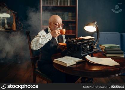Elderly writer drinks coffee at vintage typewriter in his home office. Old man in glasses writes literature novel in room with smoke, inspiration. Elderly writer drinks coffee at vintage typewriter