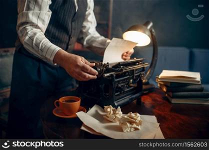 Elderly writer inserts paper into the vintage typewriter in his home office. Old man writes literature novel in room with smoke, inspiration, coffee and crumpled sheets on the table. Elderly writer inserts paper into the typewriter