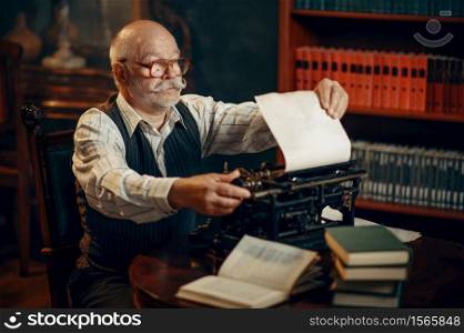 Elderly writer inserts paper into the vintage typewriter in his home office. Old man writes literature novel in room with smoke. Elderly writer inserts paper into the typewriter