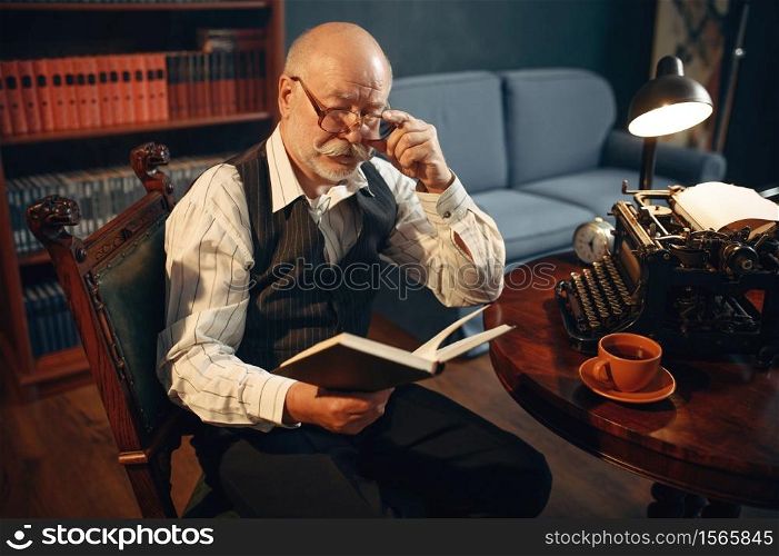 Elderly writer reads his work at vintage typewriter in home office. Old man in glasses writes literature novel in room with smoke, inspiration. Elderly writer reads work at vintage typewriter