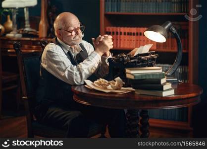 Elderly writer thinks at vintage typewriter in home office. Old man in glasses writes literature novel in room with smoke. Elderly writer thinks at vintage typewriter