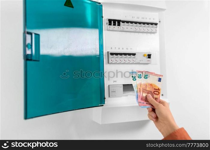electricity, energy crisis and power consumption concept - close up of hand with money and electric board at home. hand with money and electric board at home