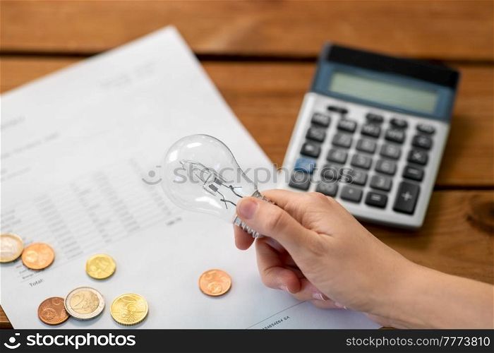 electricity, energy crisis and power consumption concept - close up of hand holding lightbulb, utility bill, calculator and money on wooden table. hand with lightbulb, bill, calculator and money