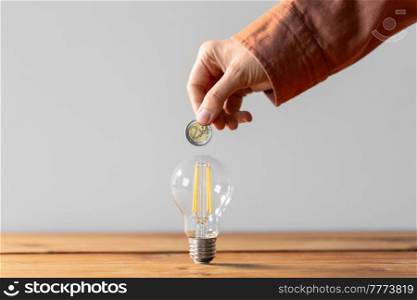 electricity, energy crisis and power consumption concept - close up of hand with coin and lightbulb on wooden table. close up of hand with coin and lightbulb on table