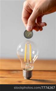 electricity, energy crisis and power consumption concept - close up of hand with coin and lightbulb on wooden table. close up of hand with coin and lightbulb on table