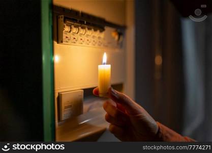 electricity, energy crisis and power consumption concept - close up of hand holding burning candle near electric board at home at night. hand with burning candle near electric board