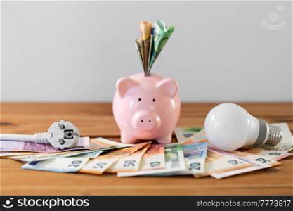 electricity, energy crisis and power consumption concept - close up of piggy bank with money, plug and lightbulb on wooden table. piggy bank with money, plug and lightbulb