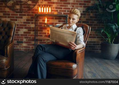 Elegance business woman in strict clothes poses on leather couch in studio, retro fashion, gangster style, mafia. Vintage lady in office with brick walls. Elegance woman in strict clothes, gangster style