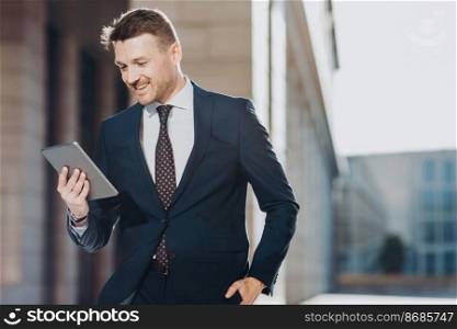 Elegant attractive male executive manager holds modern digital tablet, reads financial news on web page, rejoices having success poses outdoor, has spare time during dinner break. Job and technology