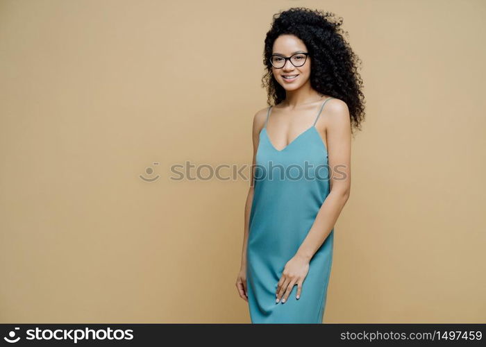 Elegant beautiful dark skinned woman with Afro hair, slim figure, wears new dress, gets ready for date rejoices new purchase stands happy indoor against beige wall copy space left for your information