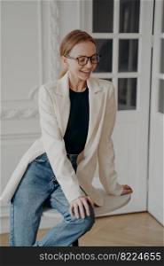 Elegant blonde european business lady in glasses and white jacket posing in studio. Confident lovely redhead woman is sitting on table. Expression of style, confidence and femininity.. Elegant blonde european business lady in glasses and white jacket posing in studio sitting on table.