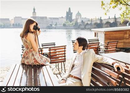 Elegant brunet young male in white formal costume poses at camera while has rest at wooden bench and admires wonderful view in front. Pretty woman uses professional camera for making photos.