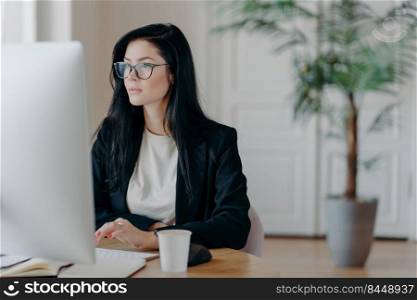 Elegant brunette businesswoman poses at cowroking space, concentrated at monitor of computer, dressed formally, drinks aromatic coffee, analyzes information, works in office, has serious expression