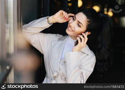 Elegant brunette slim woman with pony tail dressed in white coat having dark eyebrows, eyes, pure skin, nice manicure chatting over mobile phone touching her head with hand looking smiling aside