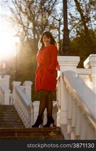 Elegant brunette woman in red coat standing on stone stairs at sunny day