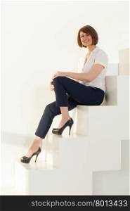 Elegant businesswoman looking at camera while sitting on stairs against of white wall. Portrait of cheerful adult businesswoman