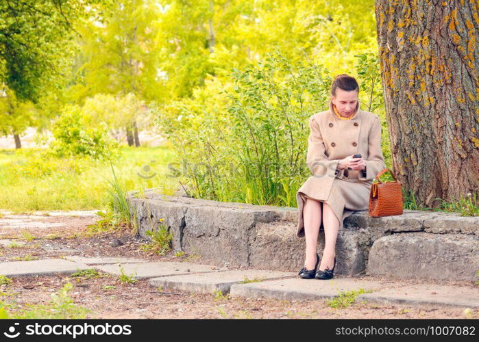 Elegant businesswoman sitting on a stone wall during a sunny spring day, and looking a message or dialing a number on her mobile phone