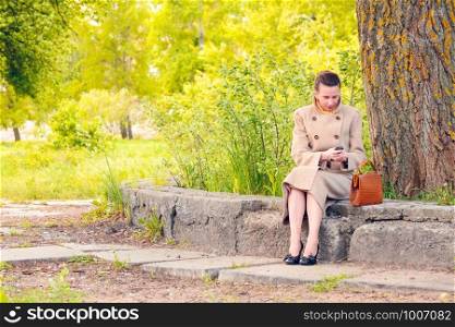 Elegant businesswoman sitting on a stone wall during a sunny spring day, and looking a message or dialing a number on her mobile phone