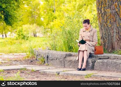 Elegant businesswoman sitting on a stone wall during a sunny spring day, and reading an adventure book