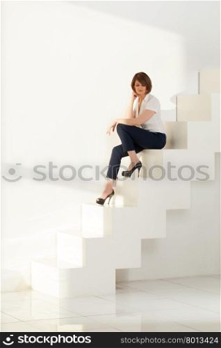 Elegant businesswoman sitting on white modern stairs against of wall in sunlight.. Adult businesswoman sitting on steps and looking down