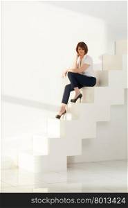 Elegant businesswoman sitting on white modern stairs against of wall in sunlight.. Adult businesswoman sitting on steps and looking down