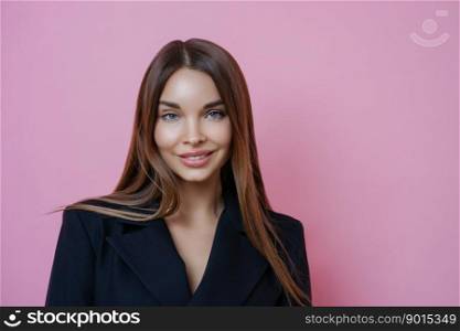 Elegant businesswoman with long straight hair, wears makeup and black formal costume, ready for business meeting, smiles pleasantly, owns her company, isolated on pink background. Indoor shot