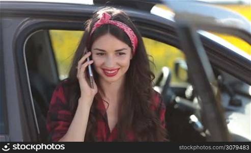 Elegant charming brunette woman talking on smartphone while sitting in the car during summer vacation trip. Attractive young hipster female chatting on mobile phone and smiling while travelling countryside by car.