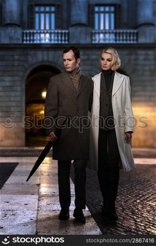 Elegant couple in coats against building facade in evening