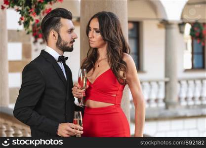 Elegant couple of lovers with a glass of wine or champagne standing on a balcony during luxury party.