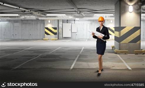 Elegant engineer woman. Young woman in hardhat with documents at underground parking. Mixed media