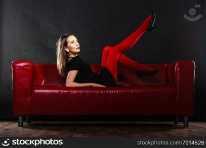 Elegant fashion outfit. Fashionable woman long legs in red vivid color pantyhose posing on couch indoor on black