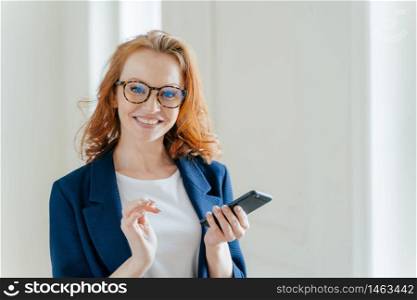 Elegant female copywriter with broad smile searches information for article and publication on cell phone wears spectacles. Businesswoman learns details of business contract online on finance platform