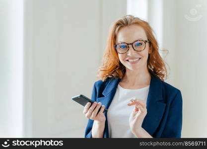 Elegant female copywriter with broad smile searches information for article and publication on cell phone wears spectacles. Businesswoman learns details of business contract online on finance platform