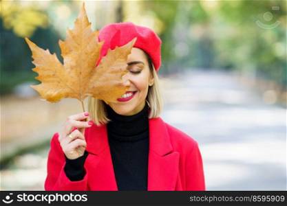 Elegant female with makeup in bright red beret and coat covering eye with autumn maple leaf while standing on walkway in park. Smiling woman covering face with dry leaf