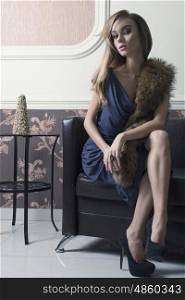 elegant female with rich fashion look is sitting on leather couch with evening blue dress, fur on shoulders and heels. Long hair and make-up.