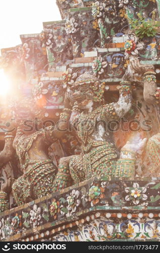 Elegant giant guardian statues with ceramic mosaic tiles decorated in ancient pagoda of Wat Arun (Temple of Dawn), glowing sun rising in the background, famous place in Bangkok, Thailand.