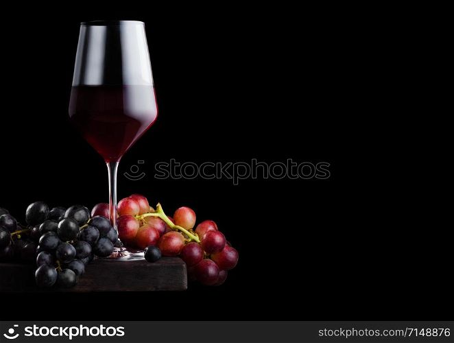 Elegant glass of red wine with dark and red grapes on wooden board on black background.