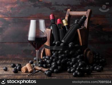 Elegant glass of red wine with dark grapes and mini bottles of wine inside vintage wooden barrel on dark wooden background. Wine still life concept.