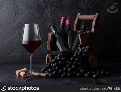 Elegant glass of red wine with dark grapes and mini bottles of wine inside vintage wooden barrel on black stone background.