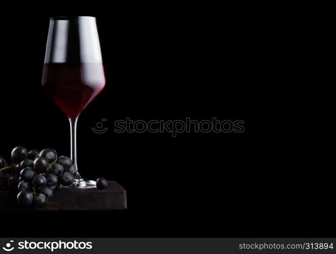 Elegant glass of red wine with dark grapes on wooden board on black background.