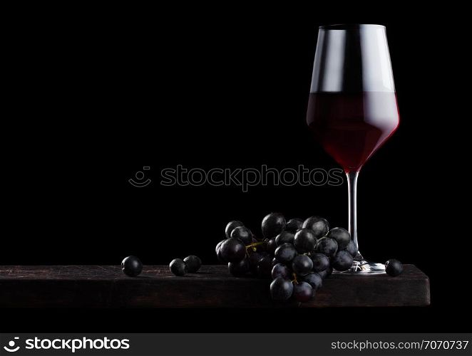 Elegant glass of red wine with dark grapes on wooden board on black background.