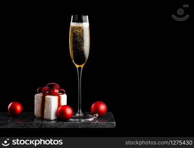 Elegant glass of yellow champagne with bubbles with christmas new year gift box and toys on marble board on black background