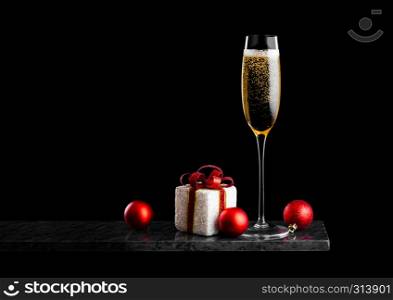 Elegant glass of yellow champagne with bubbles with christmas new year gift box and toys on marble board on black background