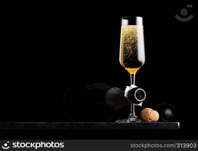 Elegant glass of yellow champagne with cork and wire cage and bottle on black marble board on black background.