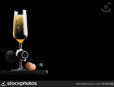 Elegant glass of yellow champagne with cork and wire cage and bottle on black marble board on black background.