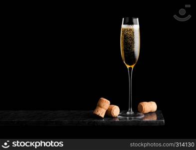 Elegant glass of yellow champagne with corks on black marble board on black.