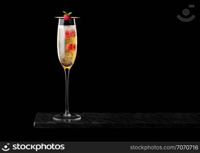 Elegant glass of yellow champagne with raspberries and bubbles inside black marble board on black background.