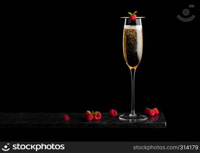 Elegant glass of yellow champagne with raspberry and fresh berries with mint leaf on stick on black marble board on black.