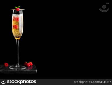 Elegant glass of yellow champagne with rasspberies and bubbles inside berries with mint leaf on stick on black marble board on black.