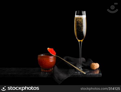 Elegant glass of yellow champagne with red caviar on golden spoon and glass container of caviar on marble board on black background